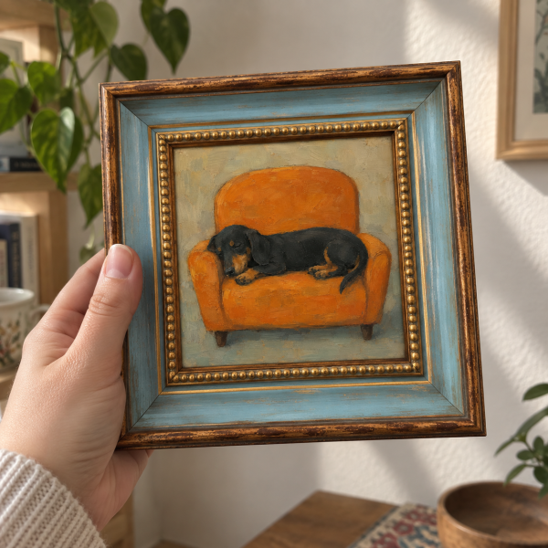 Framed oil painting of a dachshund sleeping on an orange armchair, held in a hand.